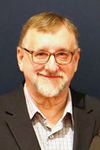 Dave Billy with glasses and a beard, wearing a suit, is smiling against a dark background.