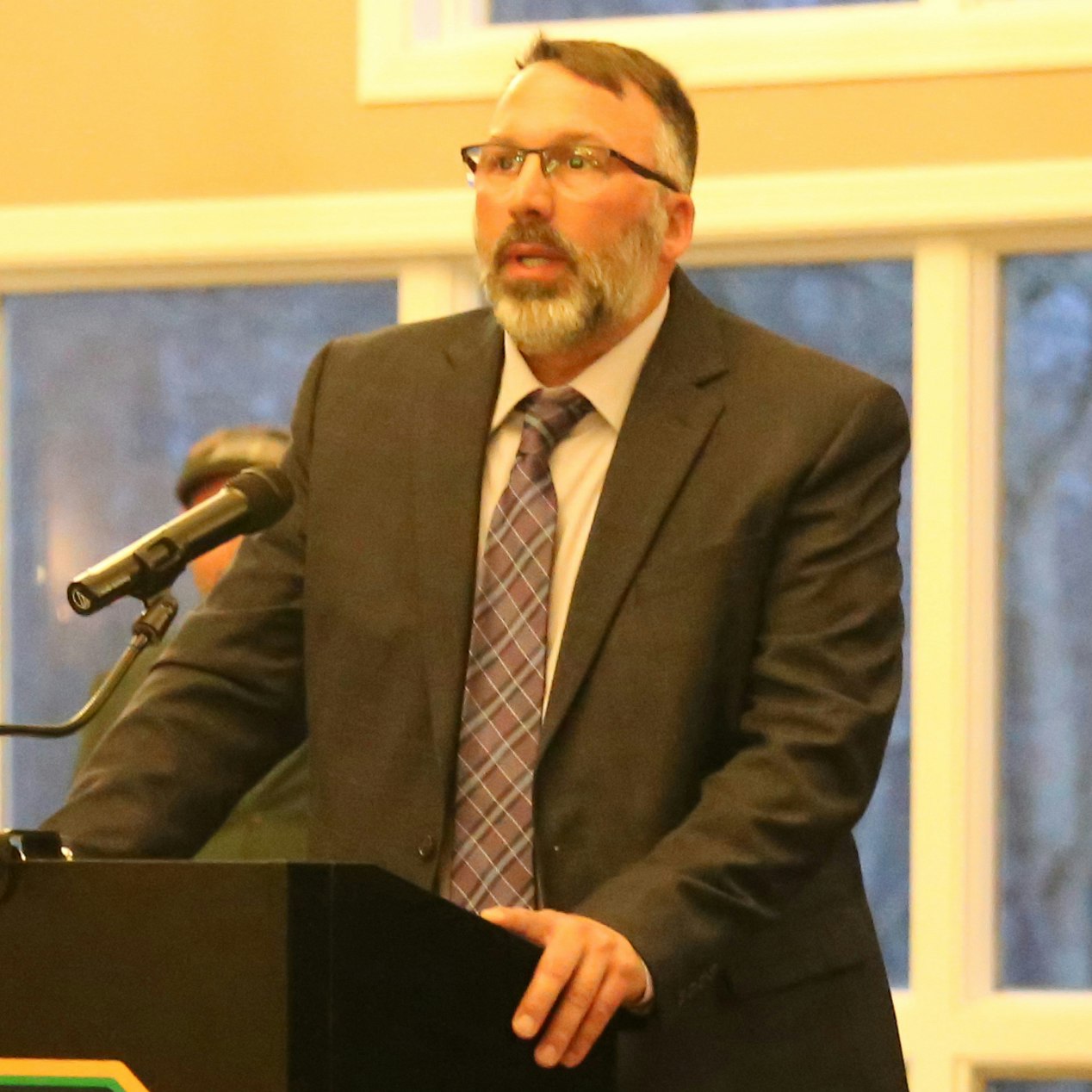 A man in a suit speaks at a podium with a microphone, likely during a formal event or presentation.