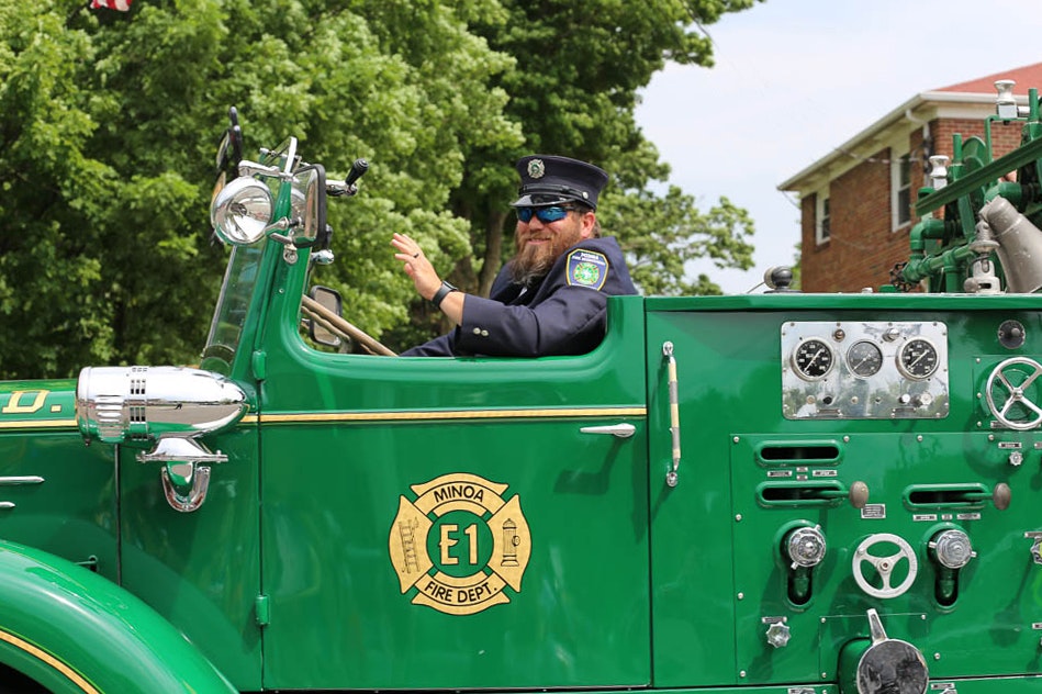 A smiling firefighter driving a vintage green fire truck, waving.