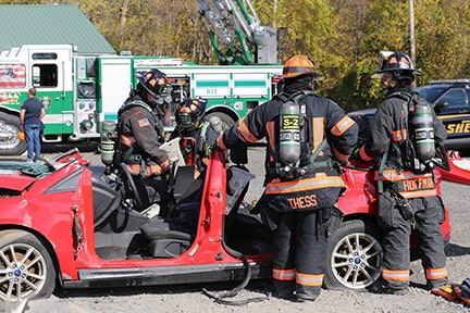 Firefighters in gear work around a red car with doors removed, possibly during an exhibition of extrication. Emergency vehicles nearby. and a crowd watching