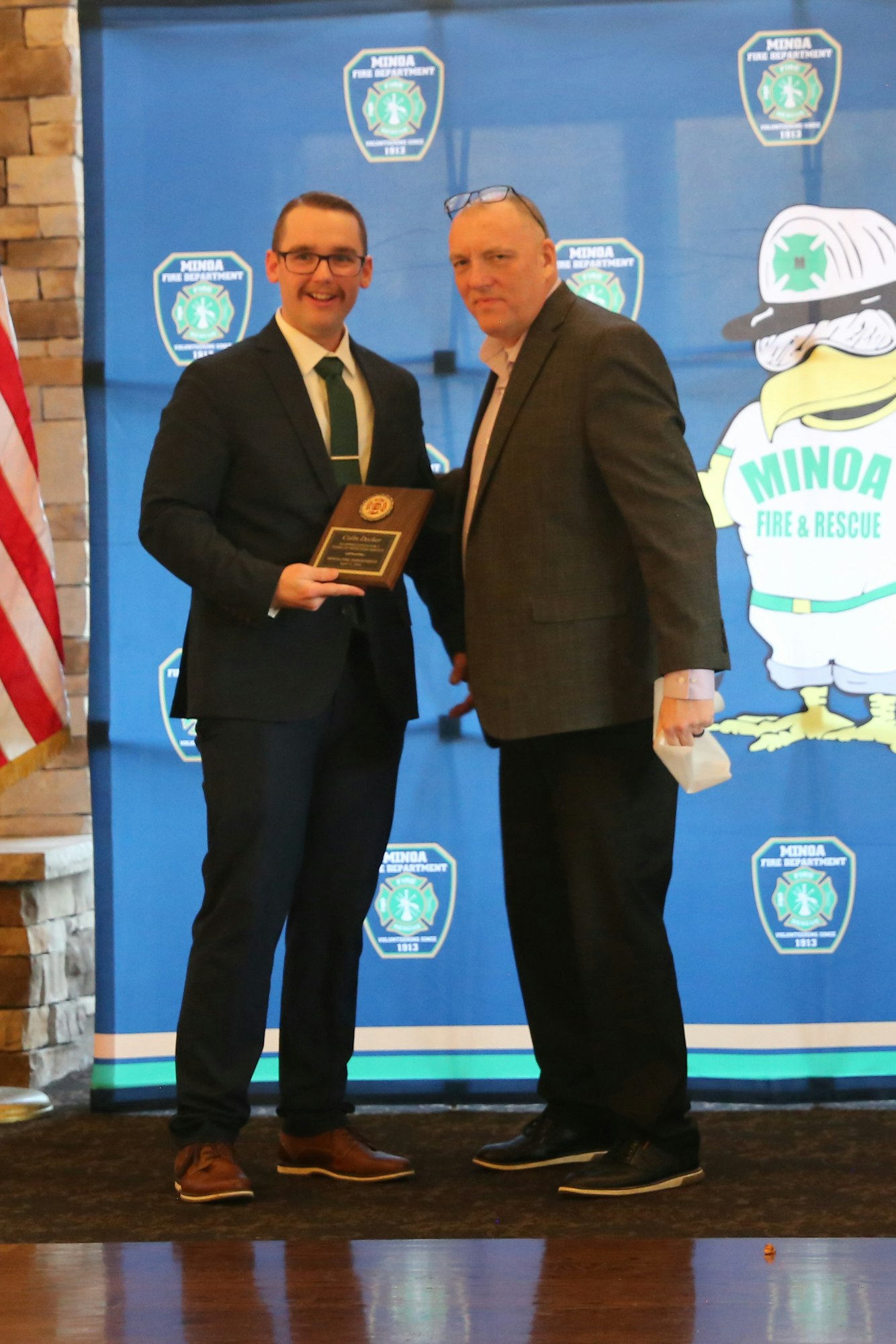 Two men are holding a plaque, smiling, in front of a backdrop for the Minoa Fire Department. An American flag is visible.