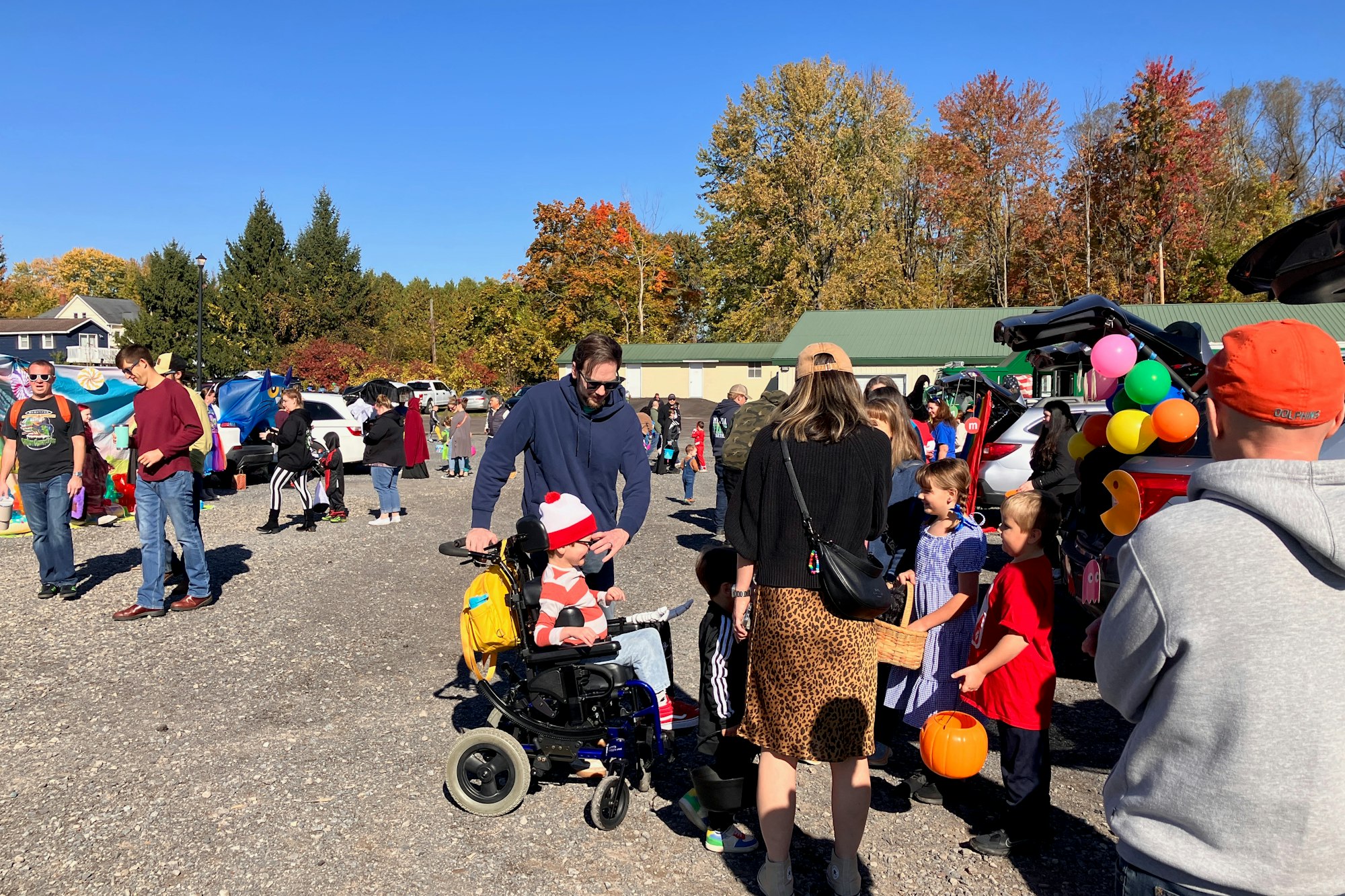 Outdoor event with people, children in costumes, colorful balloons, and cars at the trunk-or-treat.