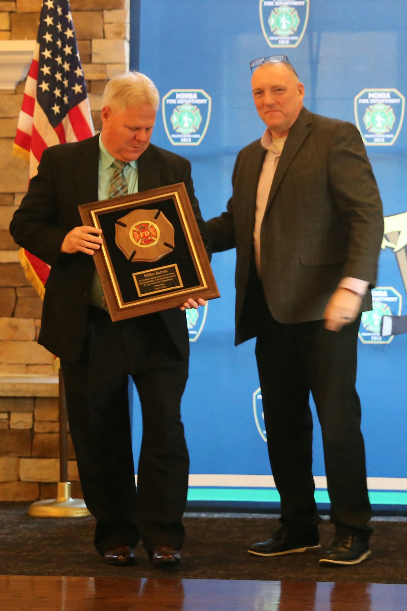 Two men at a podium are presenting an award, with an American flag in the background and a blue banner featuring a fire department emblem.