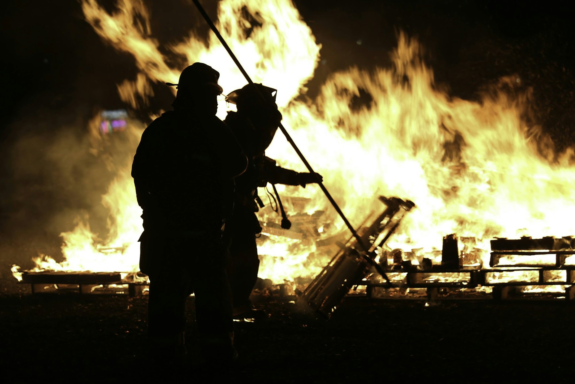 Silhouettes of firefighters battling a large blaze, surrounded by roaring flames and burning wooden pallets.