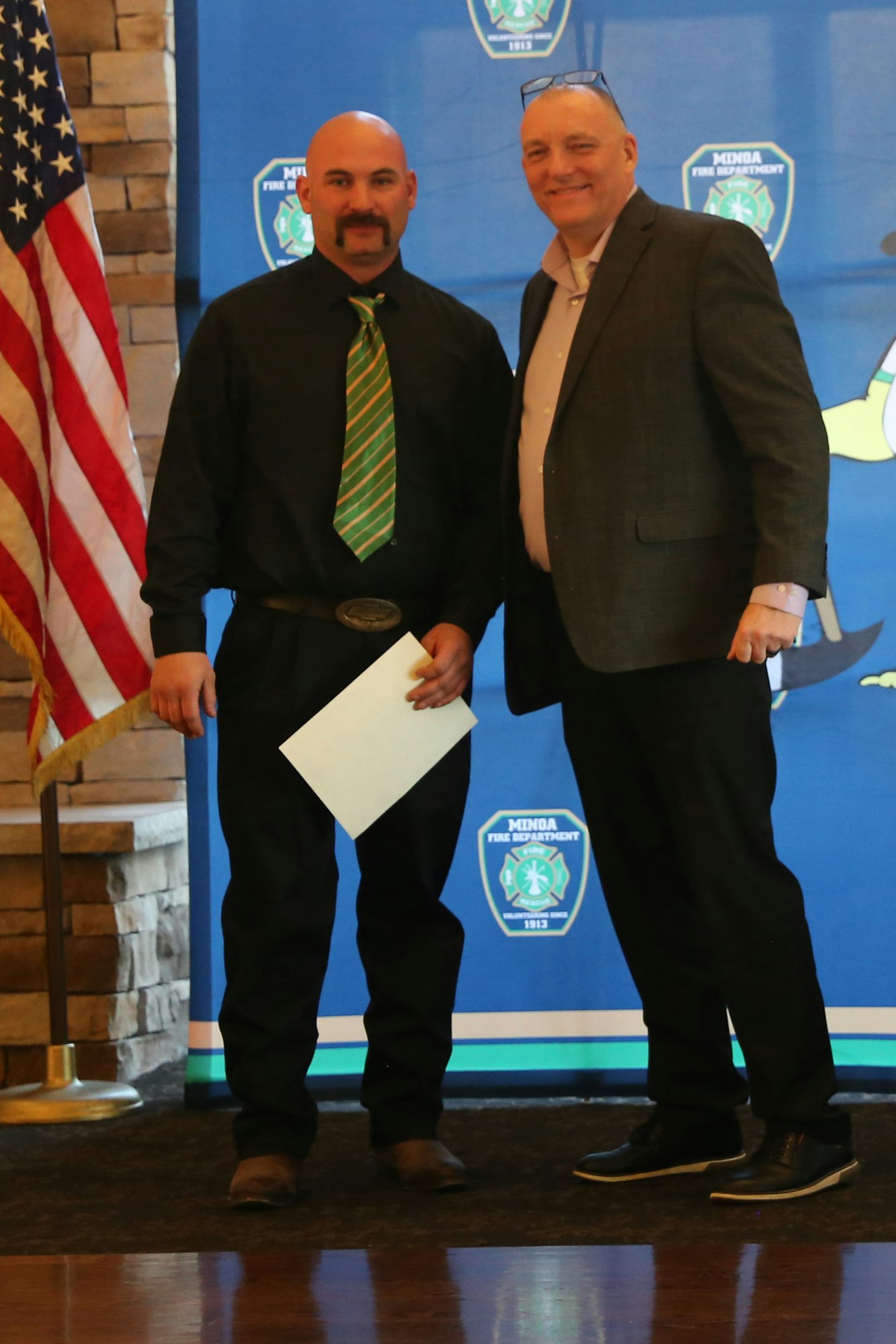 Two men stand together for a photo, one holding a certificate, with an American flag and a fire department backdrop.
