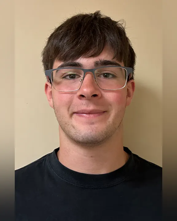 The image shows a young male with glasses, smiling, wearing a black t-shirt against a light beige background.