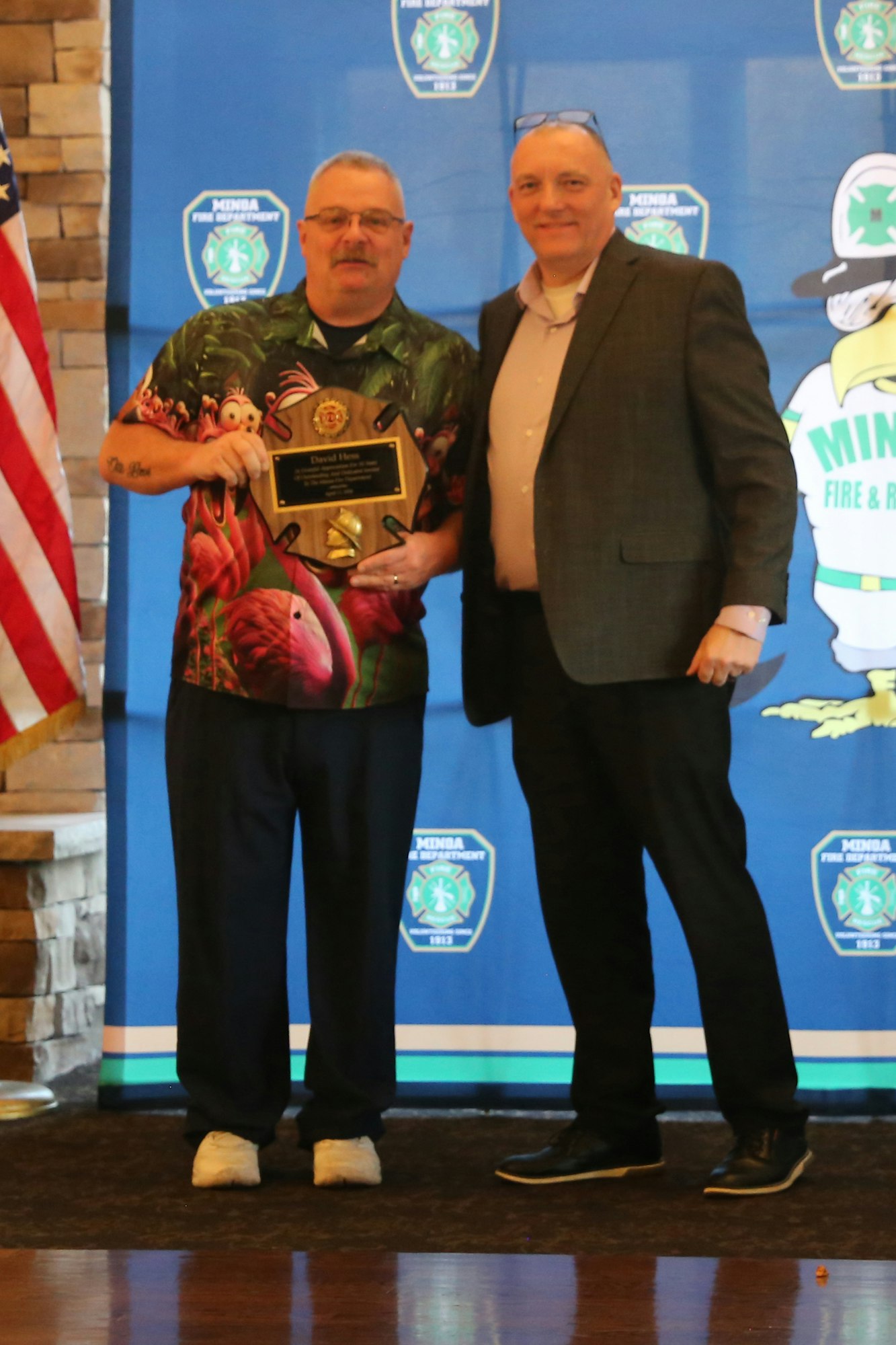 Two men stand together, one holding an award plaque, with a backdrop featuring a fire department logo and an American flag.