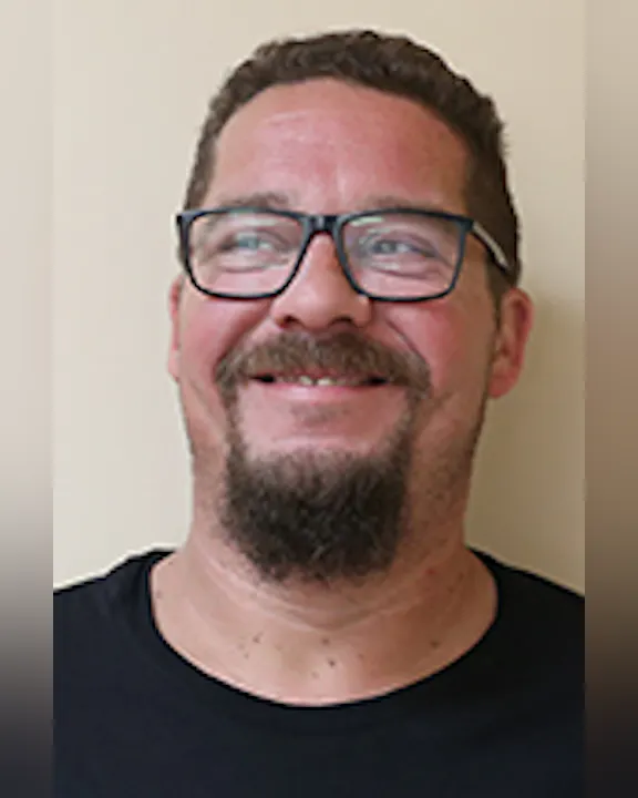 Brad Cretaro with glasses, a beard, and a black shirt smiling against a beige background.