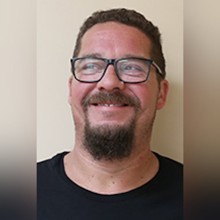 Brad Cretaro with glasses, a beard, and a black shirt smiling against a beige background.