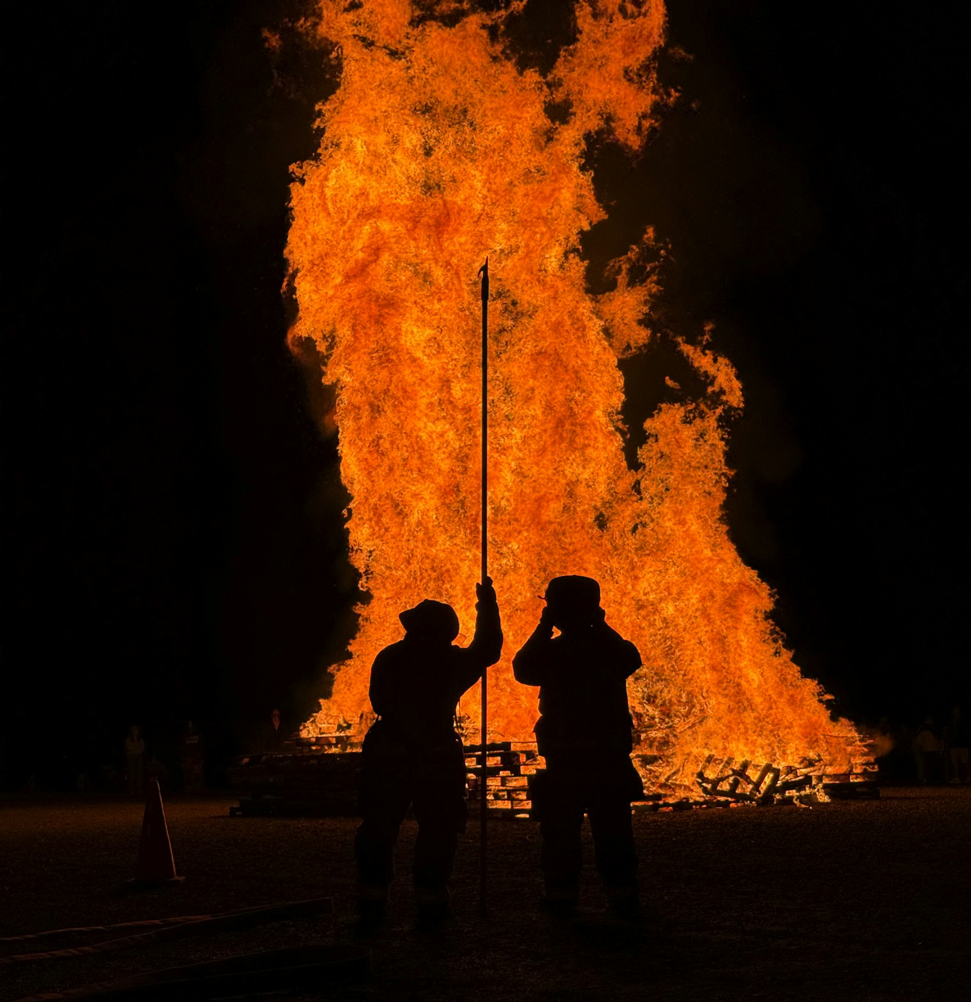 Two silhouetted Minoa firefighters in gear by a large bonfire at night, with one holding a pole.