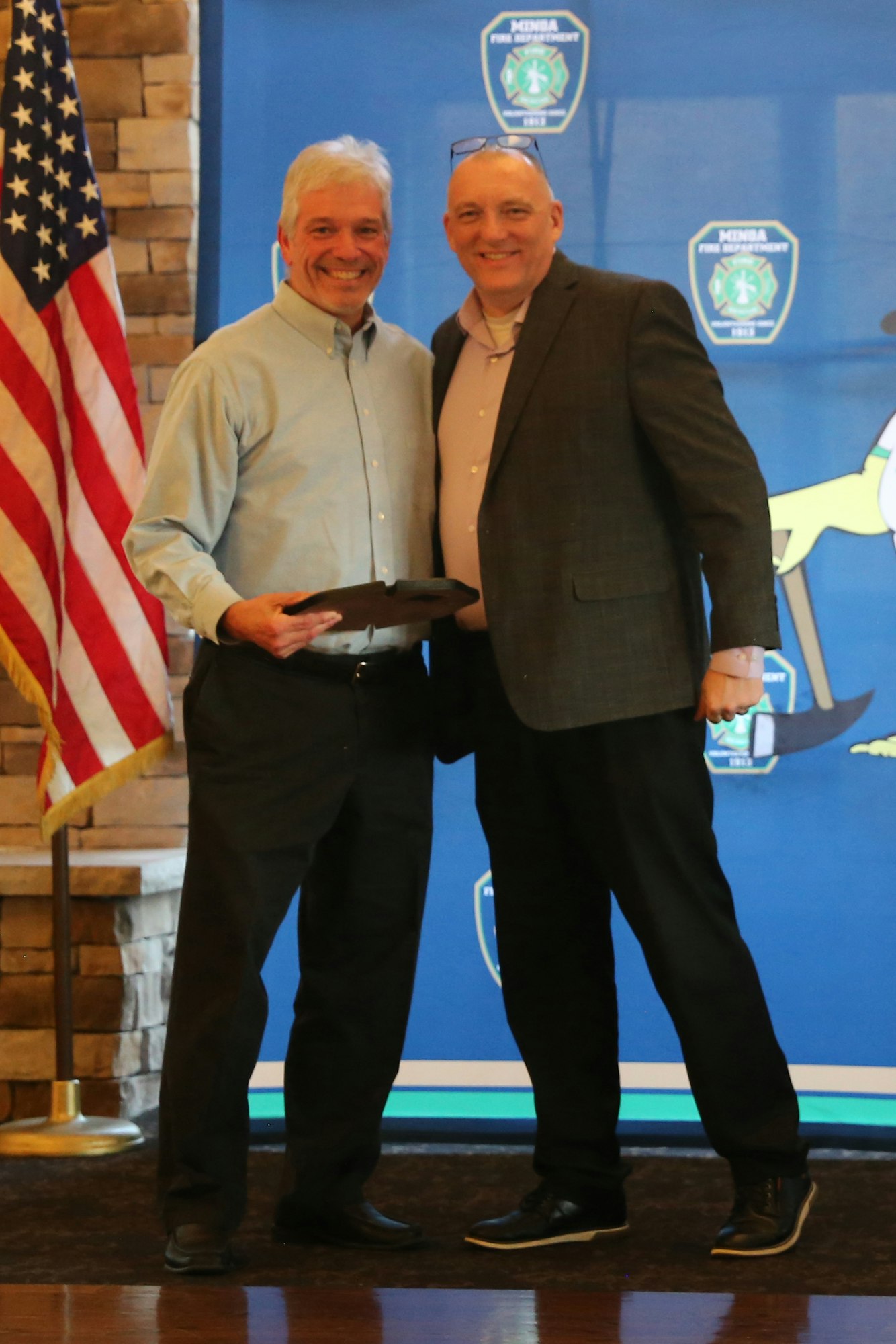 Two men are smiling and posing for a photo while holding an award, with an American flag and a backdrop featuring a shield logo.