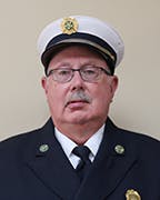 Pat Flannery in a formal uniform, likely a firefighter, with badges, glasses, and a cap, against a neutral background.