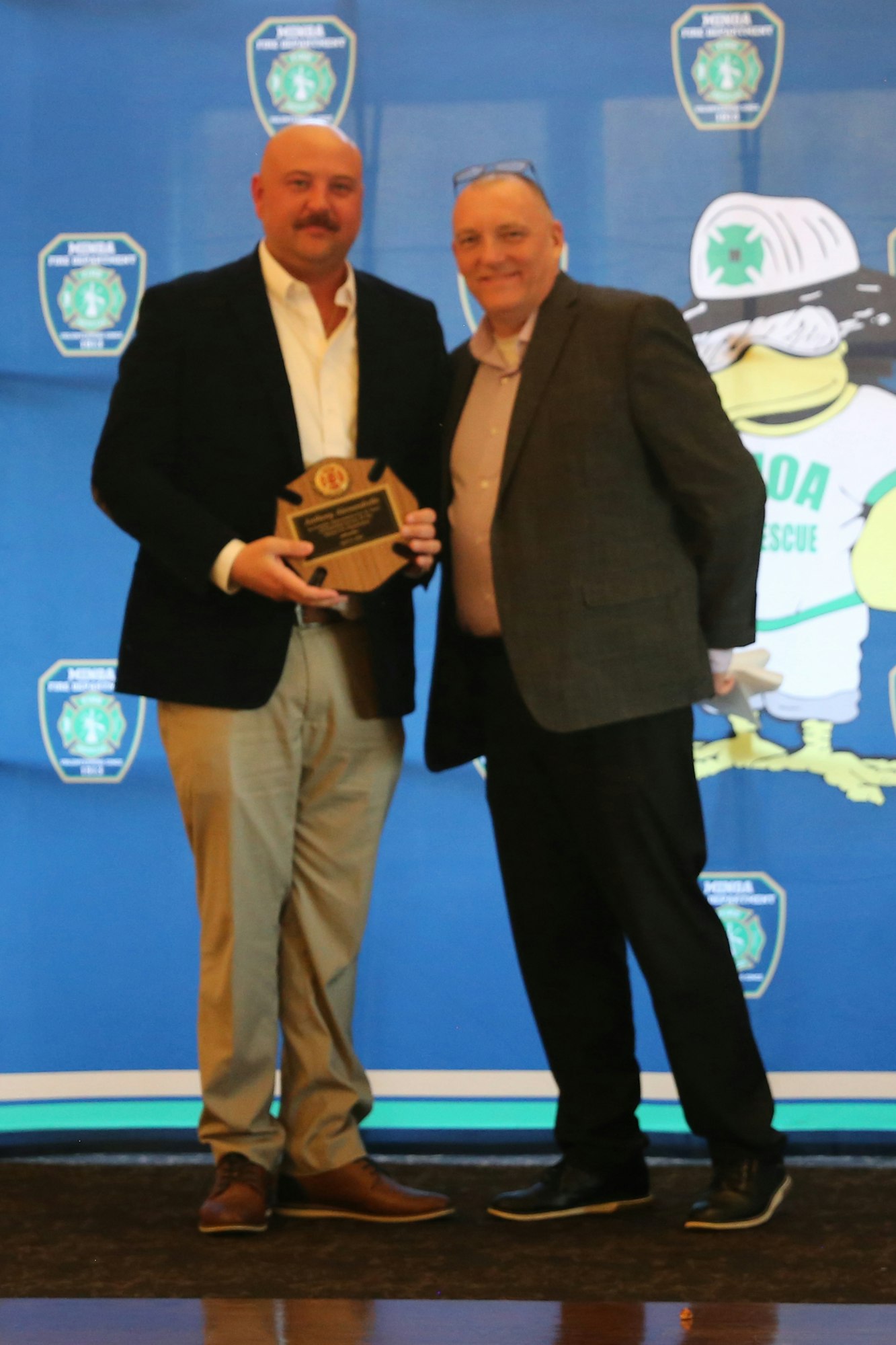 A man receives an award from another in front of a blue backdrop with emergency service logos.