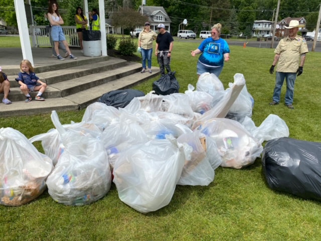 Collection of trash bags on grass, with people standing and sitting around, after the community cleanup.