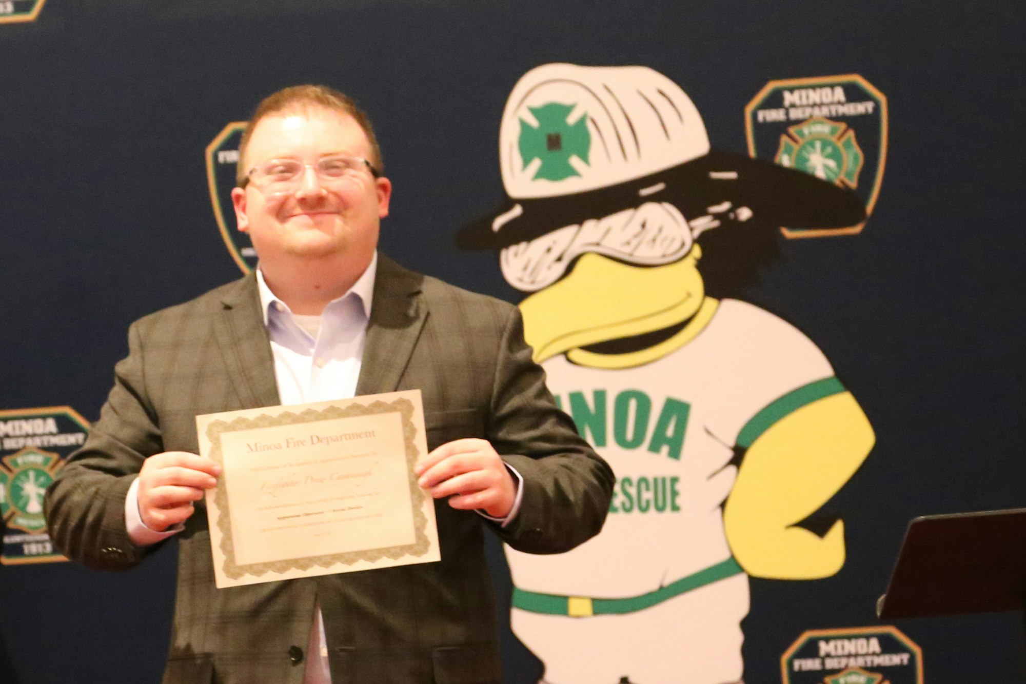 Person holding a certificate from Minoa Fire Department, standing in front of a cartoon mascot backdrop.