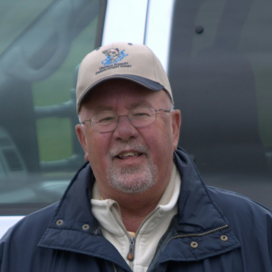 A smiling man wearing glasses and a baseball cap, dressed in a jacket, is in front of a vehicle.