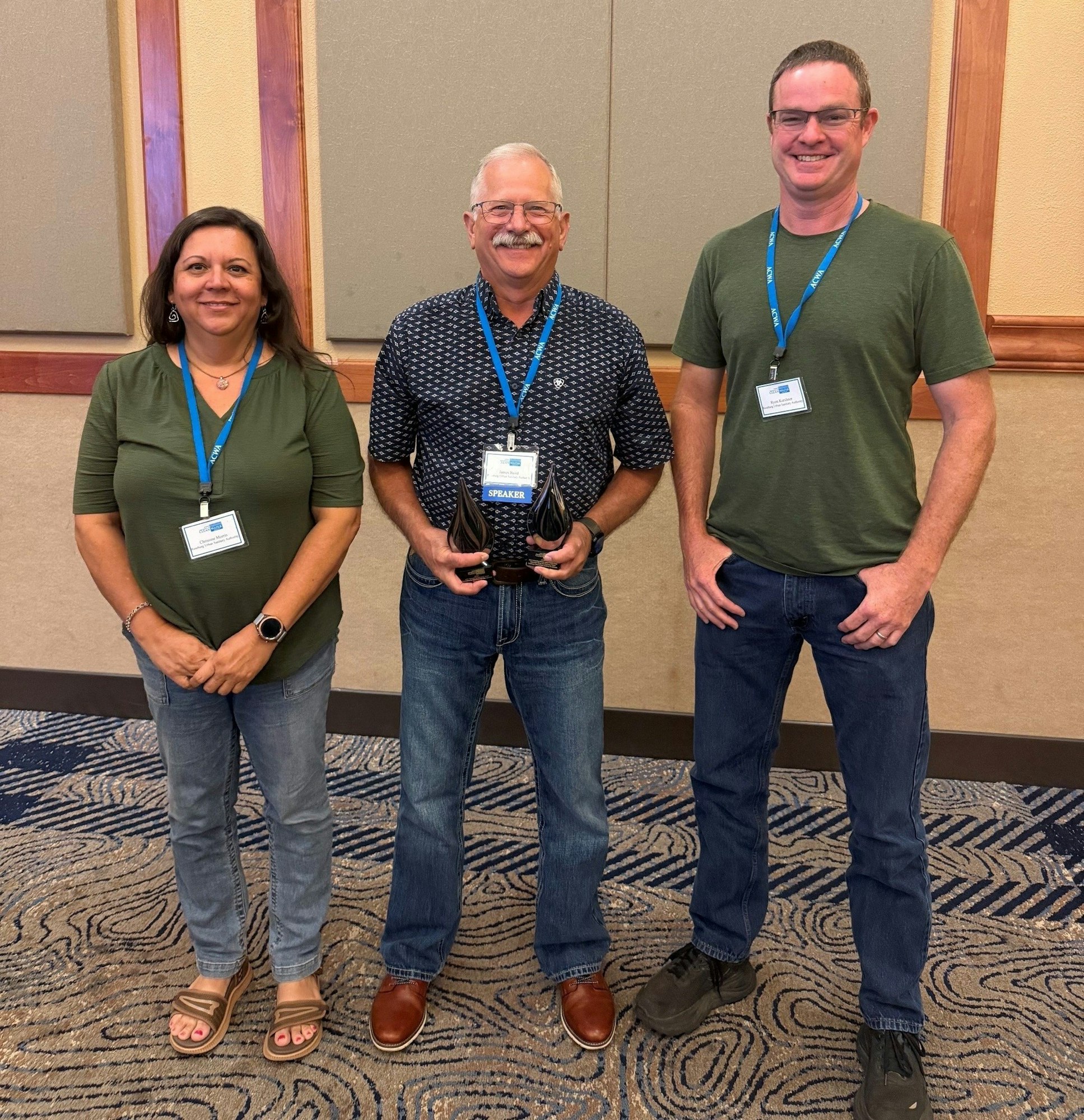 Three individuals stand together, one holding an award, smiling, and wearing name badges in a conference setting.