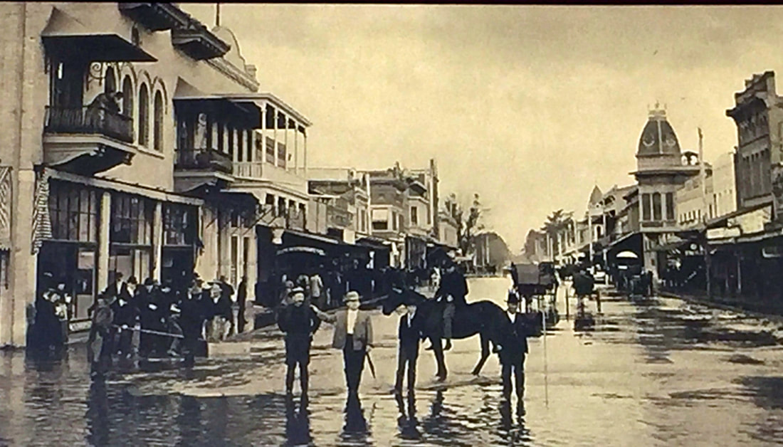Historic street scene with people in water due to flooding, horse with rider, vintage buildings, and a bustling early 20th-century atmosphere.