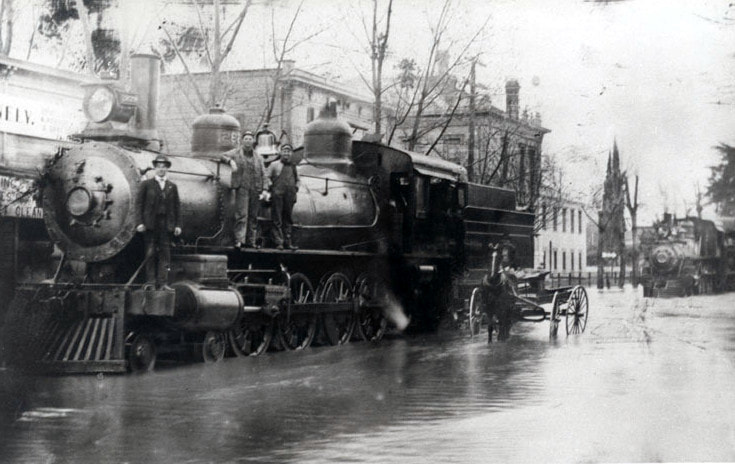 Old steam locomotive on a flooded street with people standing on it, surrounded by buildings and trees.
