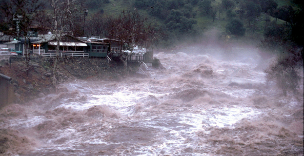 Raging floodwaters rush past a partially submerged town surrounded by trees and hills in the background.
