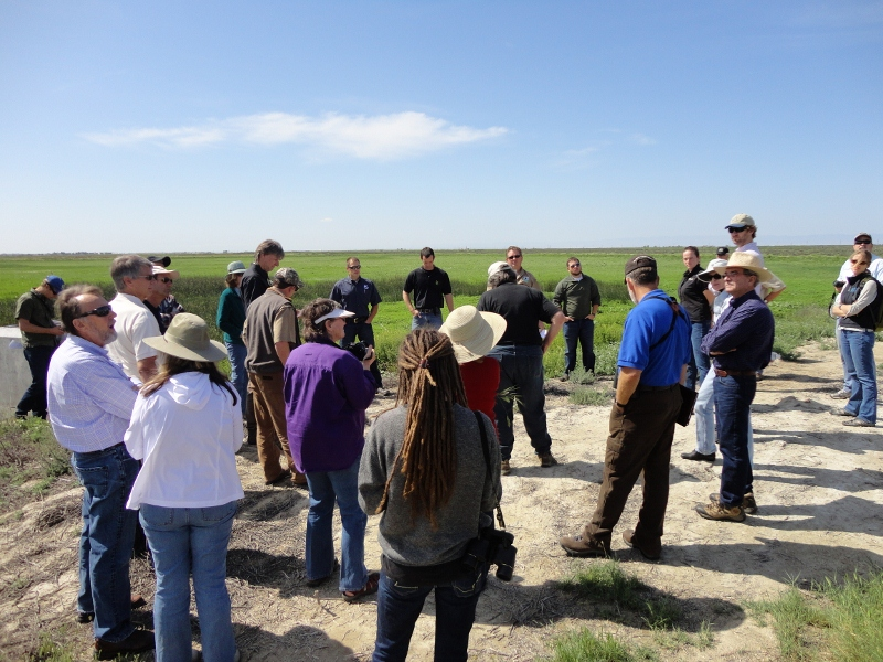 A group of people stands in a circle outdoors in a field, some wearing hats, under a clear sky.