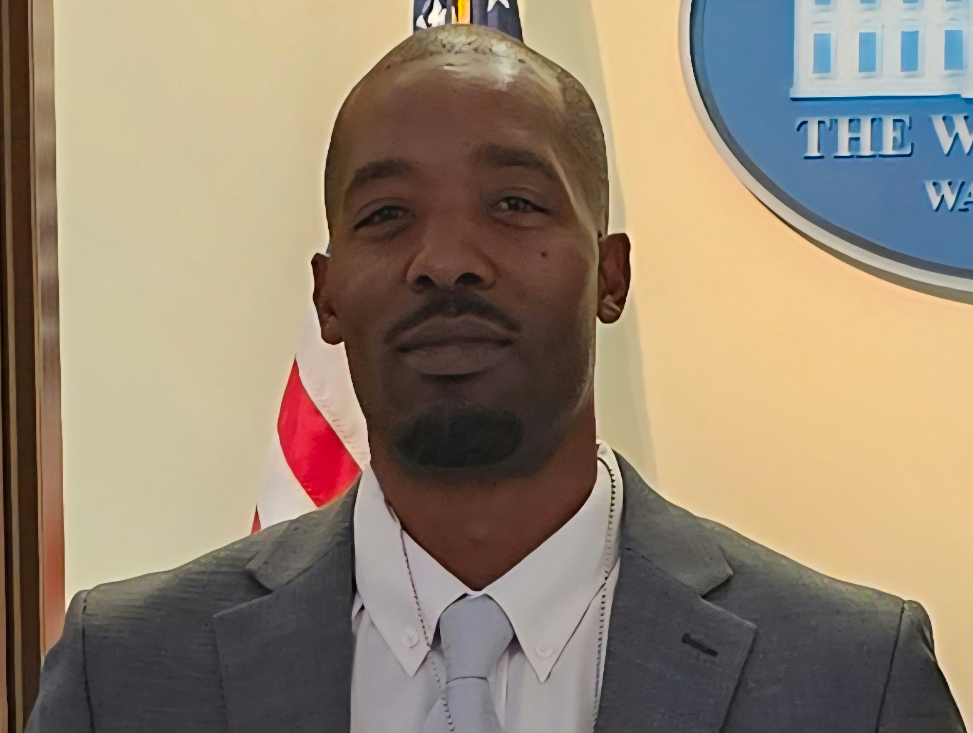 A person in a suit stands in front of a U.S. flag and a "The White House, Washington" sign.