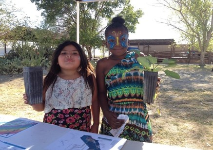 Two children holding plant seedlings, one has face paint, standing outdoors near a table with papers.
