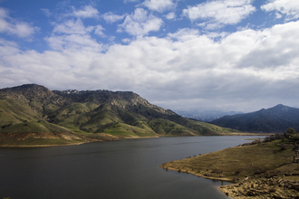 A serene lake bordered by grassy hills and mountains under a partly cloudy sky.