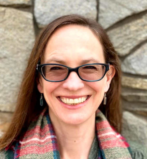 A person with glasses smiling, wearing a colorful scarf, in front of a stone wall background.