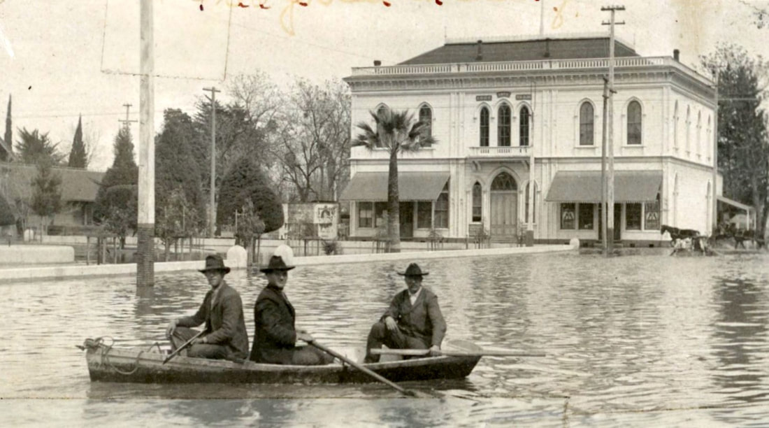 Three men in a canoe navigating a flooded street in front of a large building, with a horse-drawn carriage in the background.