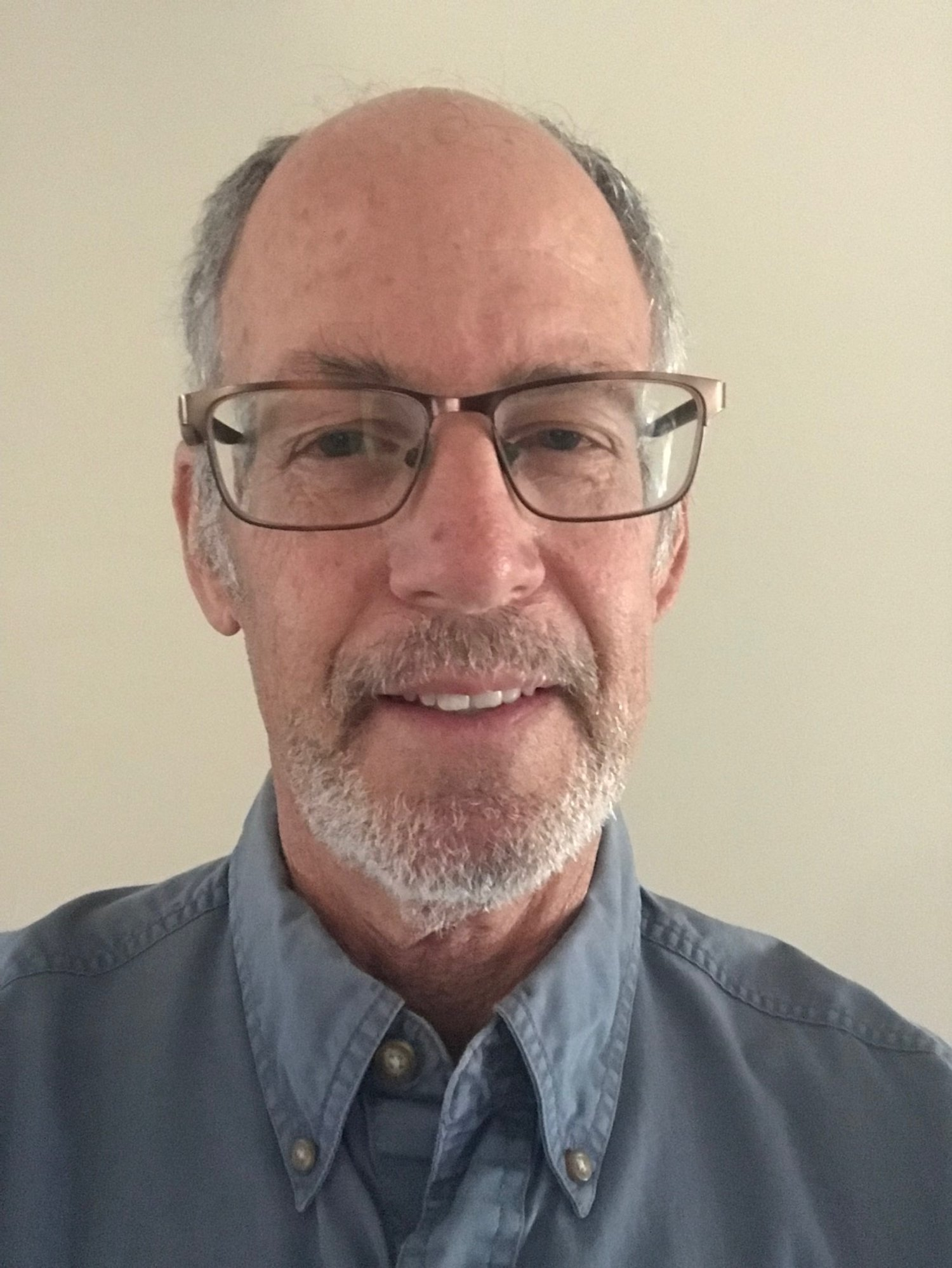 A man with glasses, a grey beard, and a button-up shirt, smiling in front of a plain background.
