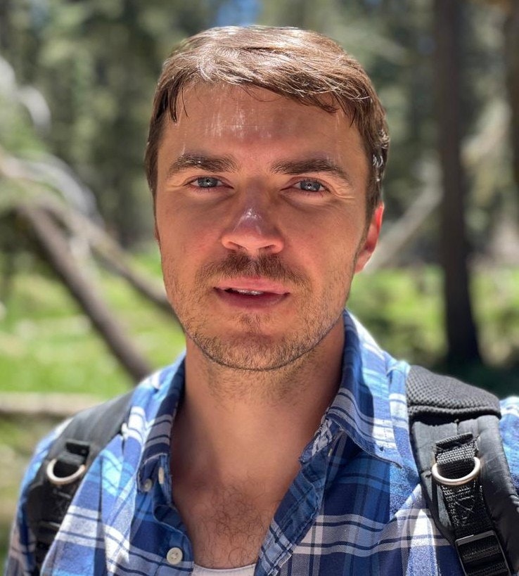 A man in a blue plaid shirt and backpack stands outdoors in a forest setting.