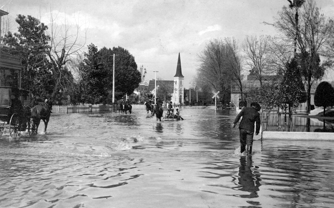 Flooded street with people, horse-drawn carriages, and a building with a steeple in the background.
