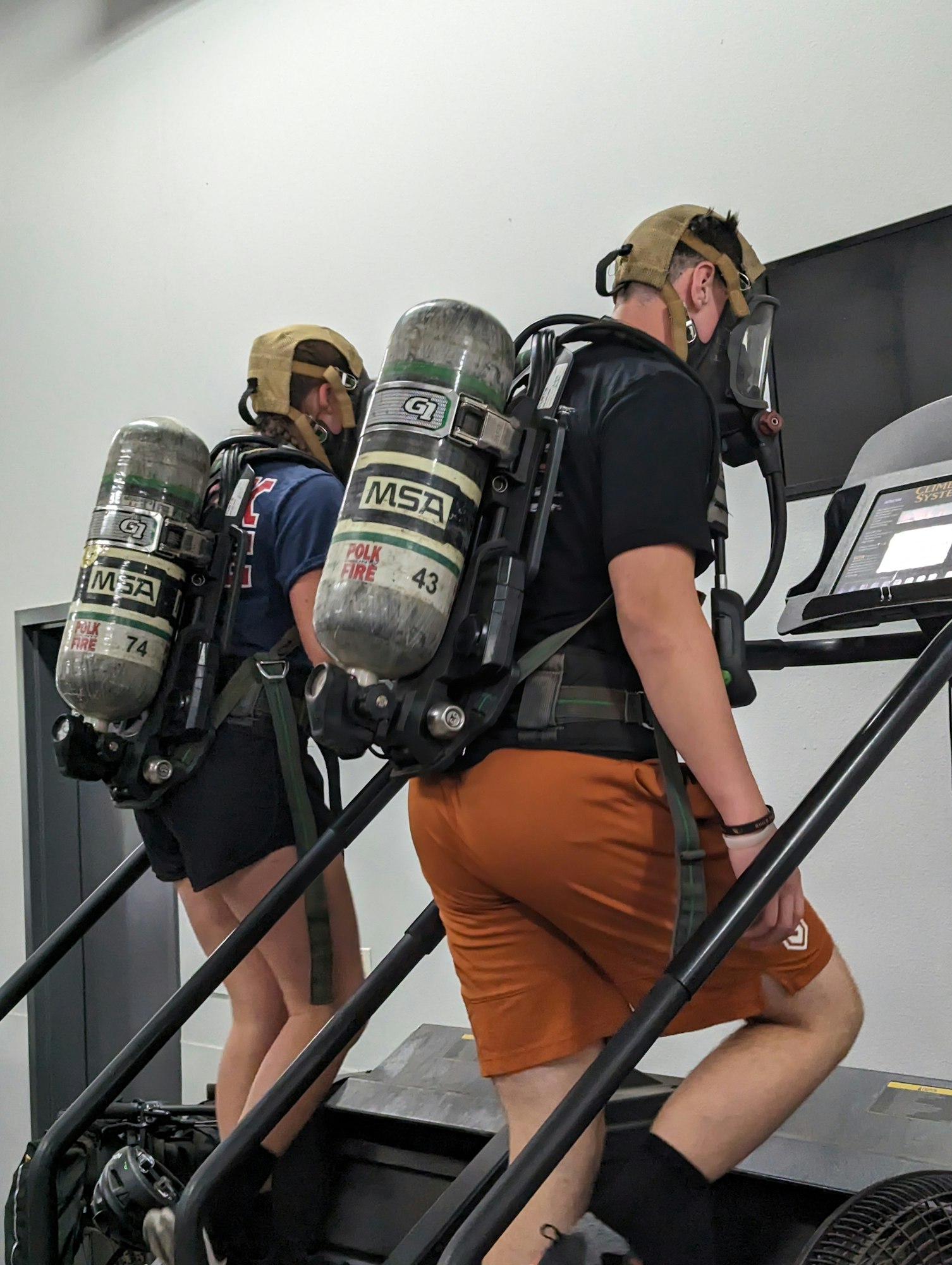 Two people with SCBA tanks on a stair machine, likely training.