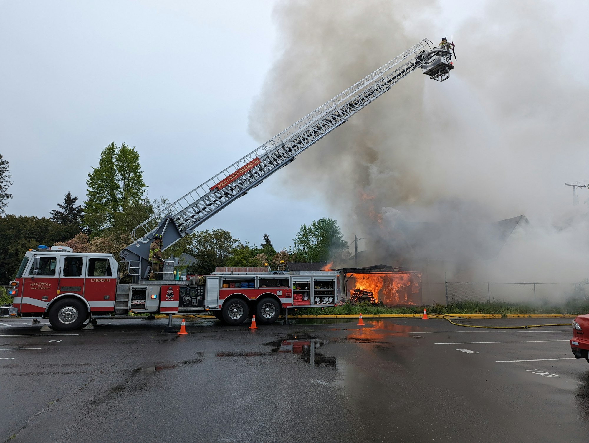 A fire truck with extended ladder fights a building fire, smoke billowing, firefighter on ladder, emergency in progress.