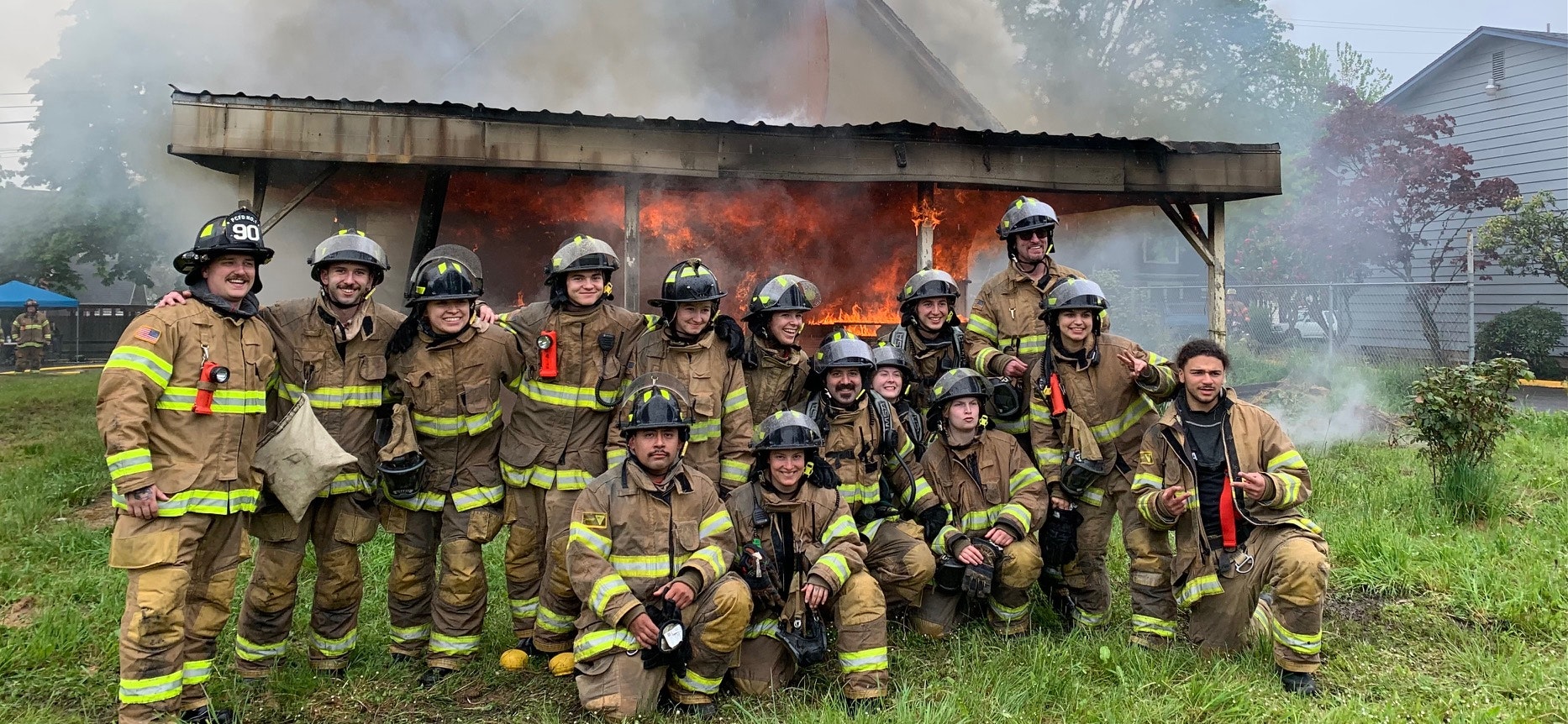 Group of firefighters in gear posing in front of a burning structure.