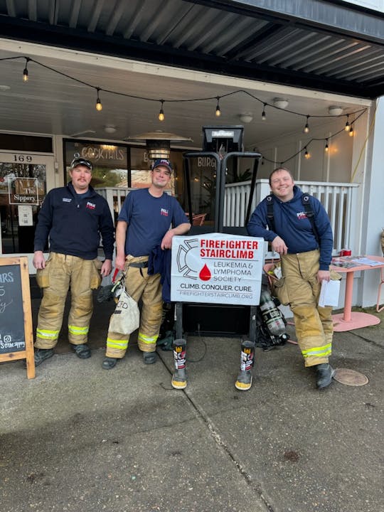 Three firefighters in gear next to a sign for a stair climb event supporting leukemia and lymphoma research.