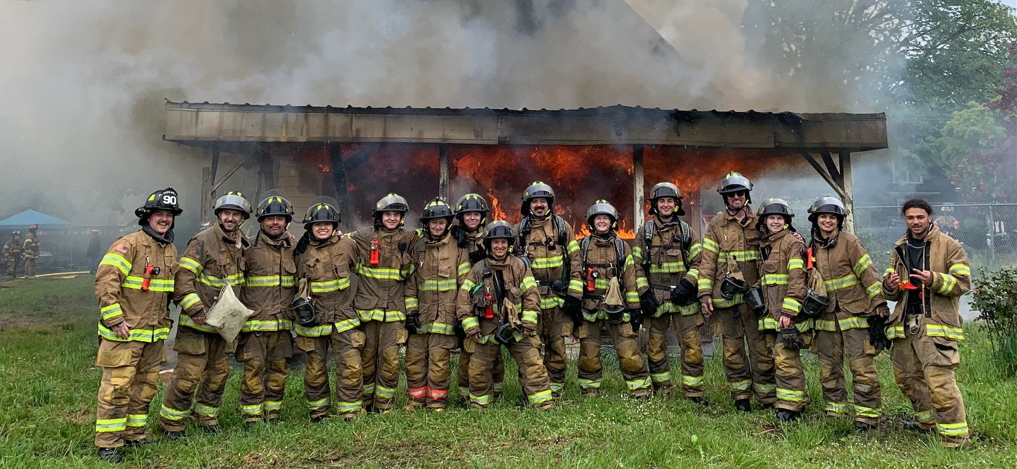 Firefighters posing in front of a burning structure, smiling, possibly during a training exercise.