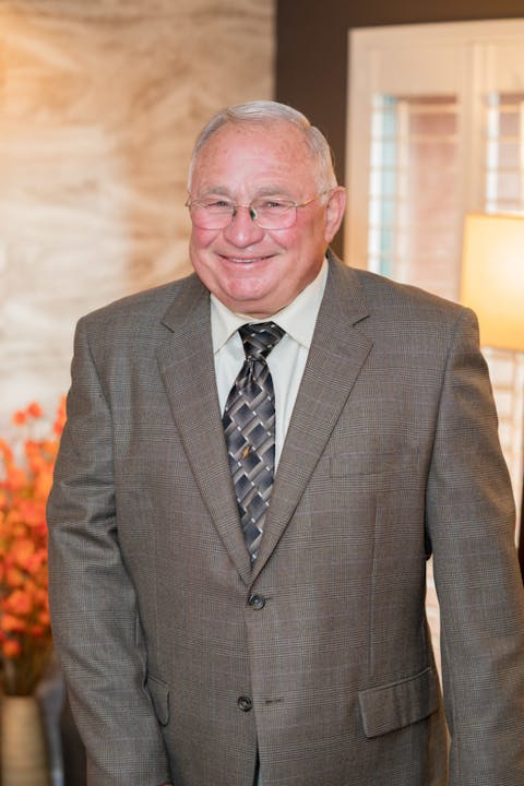 Image description: A photograph of Graig Stephens, standing and smiling. He is wearing glasses, a suit, and has white hair.