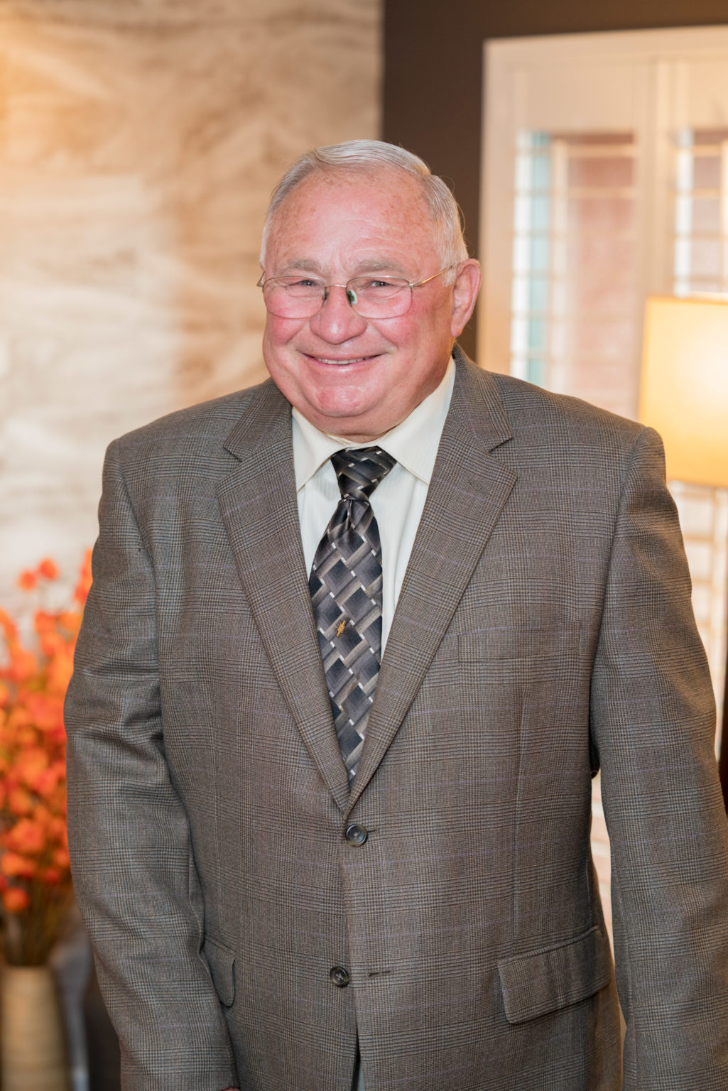 Image description: A photograph of Graig Stephens, standing and smiling. He is wearing glasses, a suit, and has white hair.