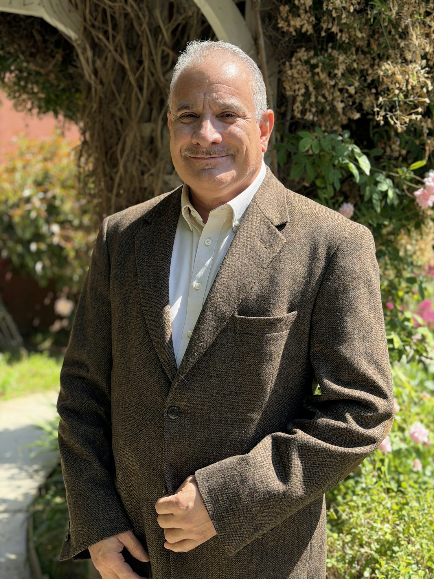 A smiling man in a brown jacket stands outdoors, surrounded by greenery and flowers.