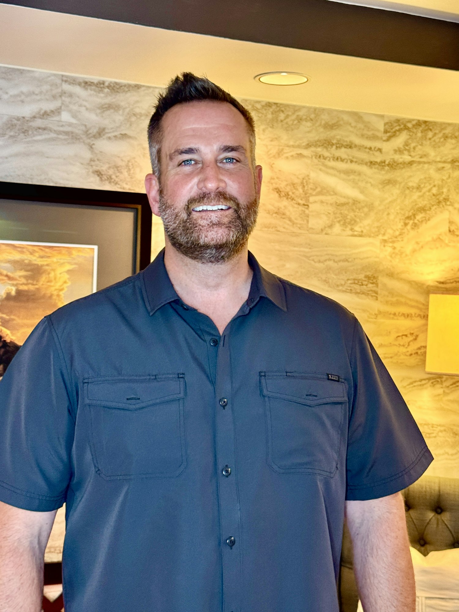 A smiling Ken Klemme in a blue shirt stands in a warmly lit room with a textured wall and framed art.