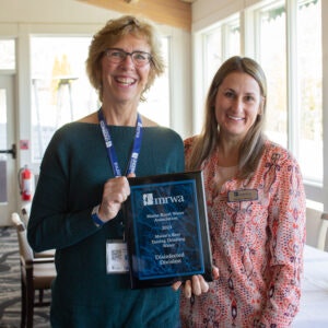 Two women are smiling together, one holding an award plaque, likely at a celebration or recognition event.