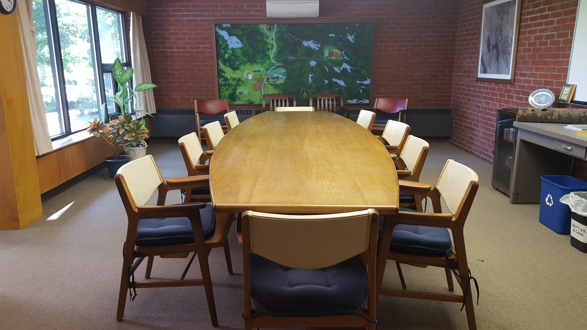 A conference room with a large wooden table, chairs arranged around it, plants, and a wall mural.