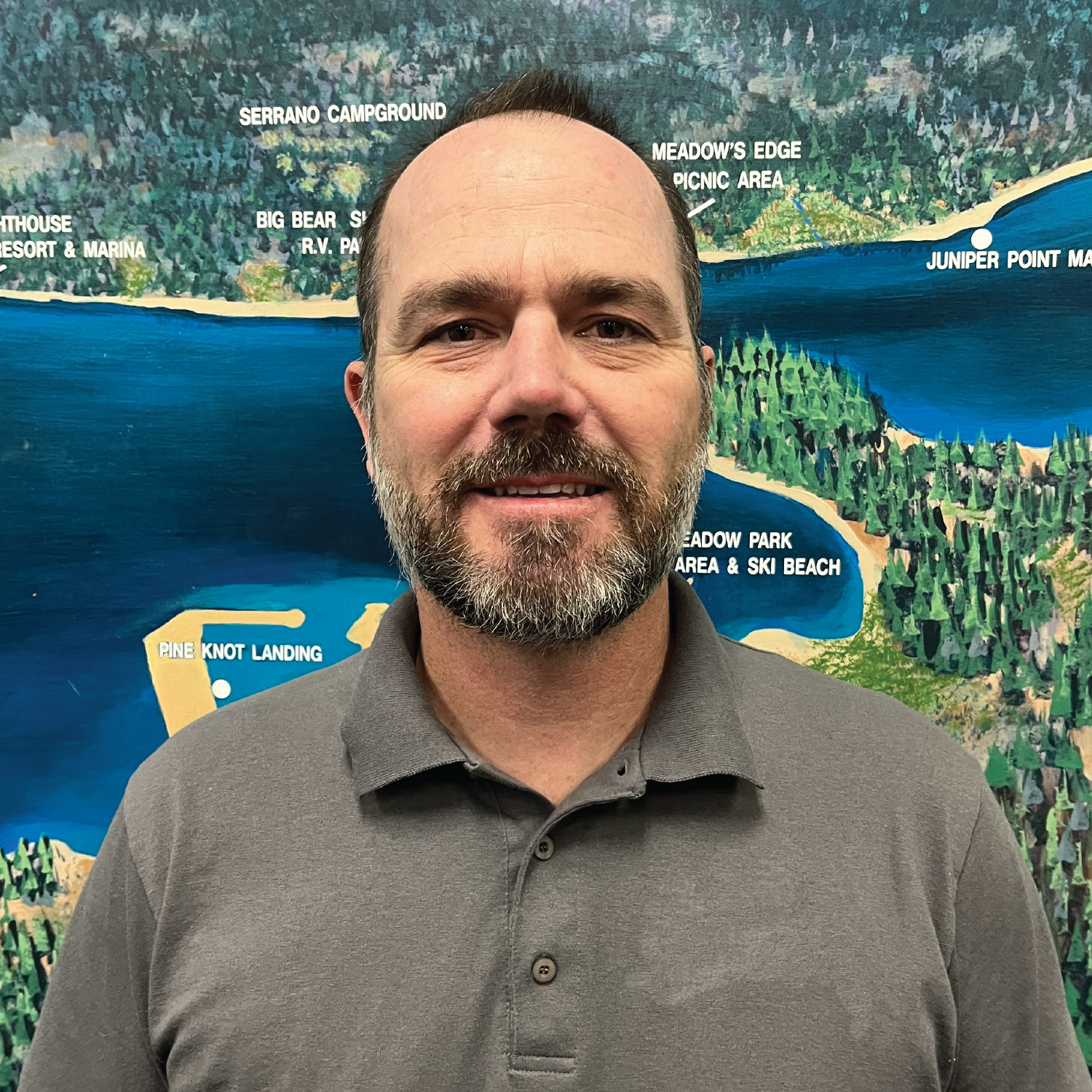 A man with a beard is smiling in front of a scenic map featuring lakes and campgrounds.