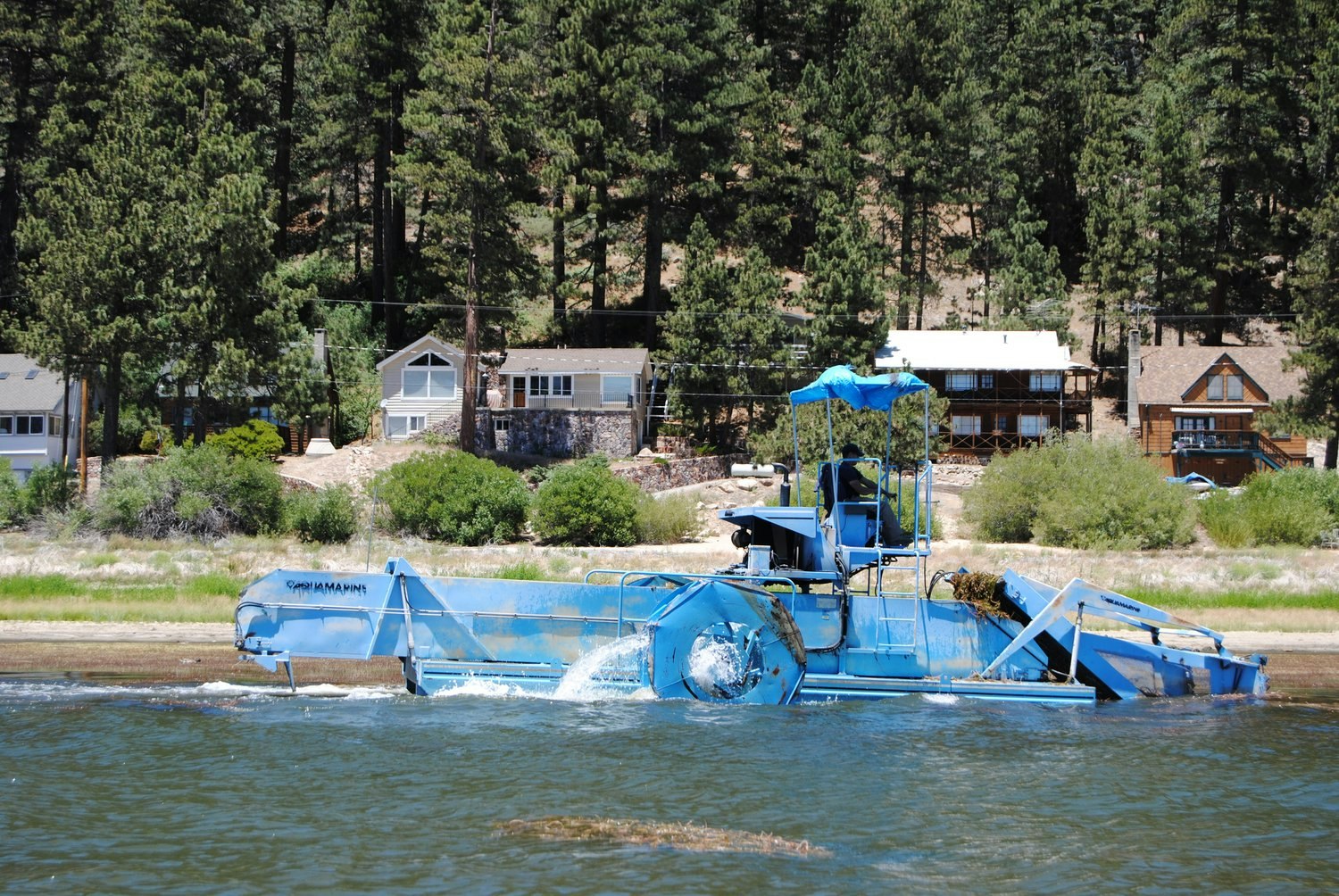 A blue aquatic weed harvester boat operating on water with houses and trees in the background.
