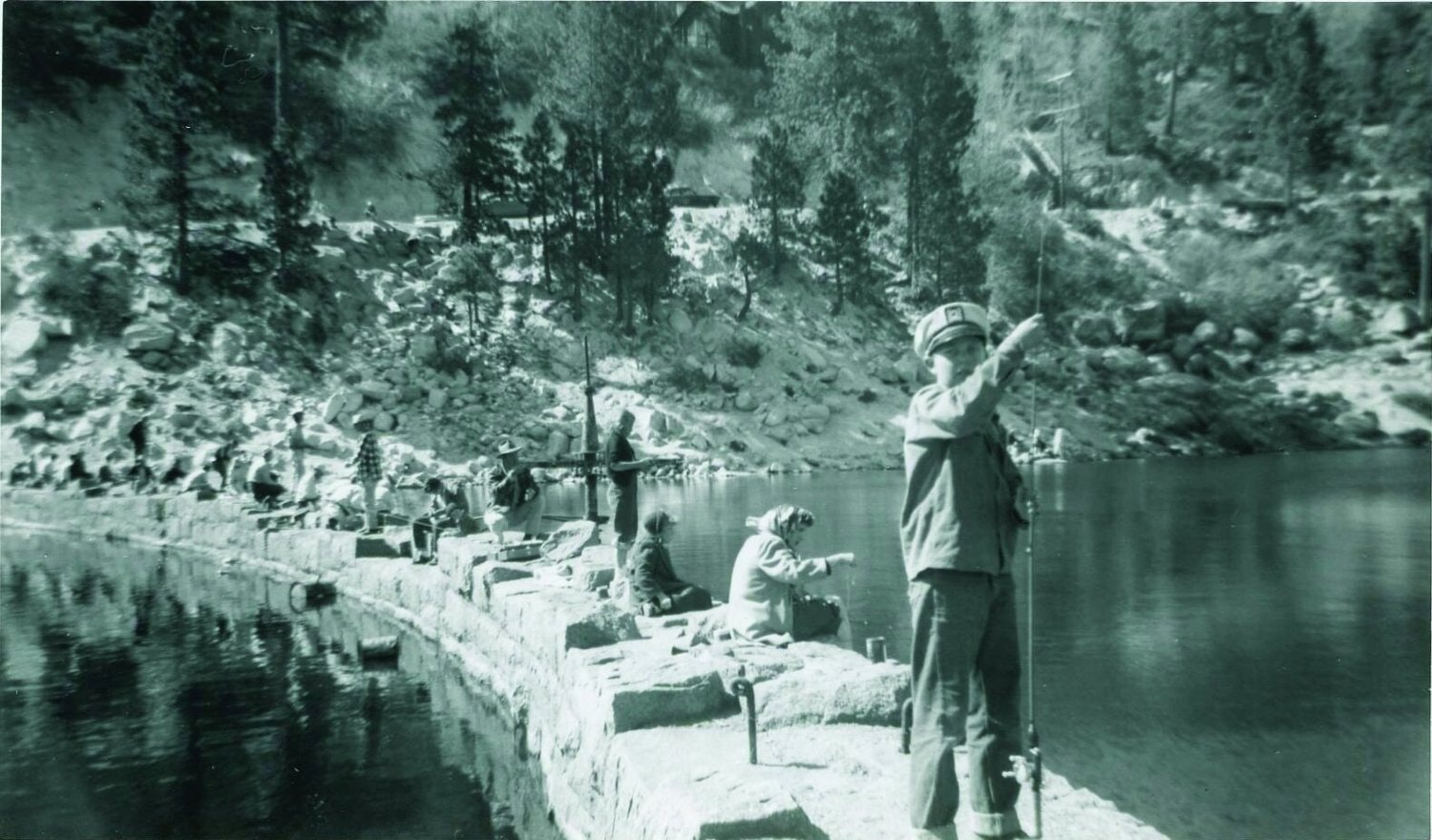 A black and white photo of people fishing by a calm lake with a wooded hill in the background.
