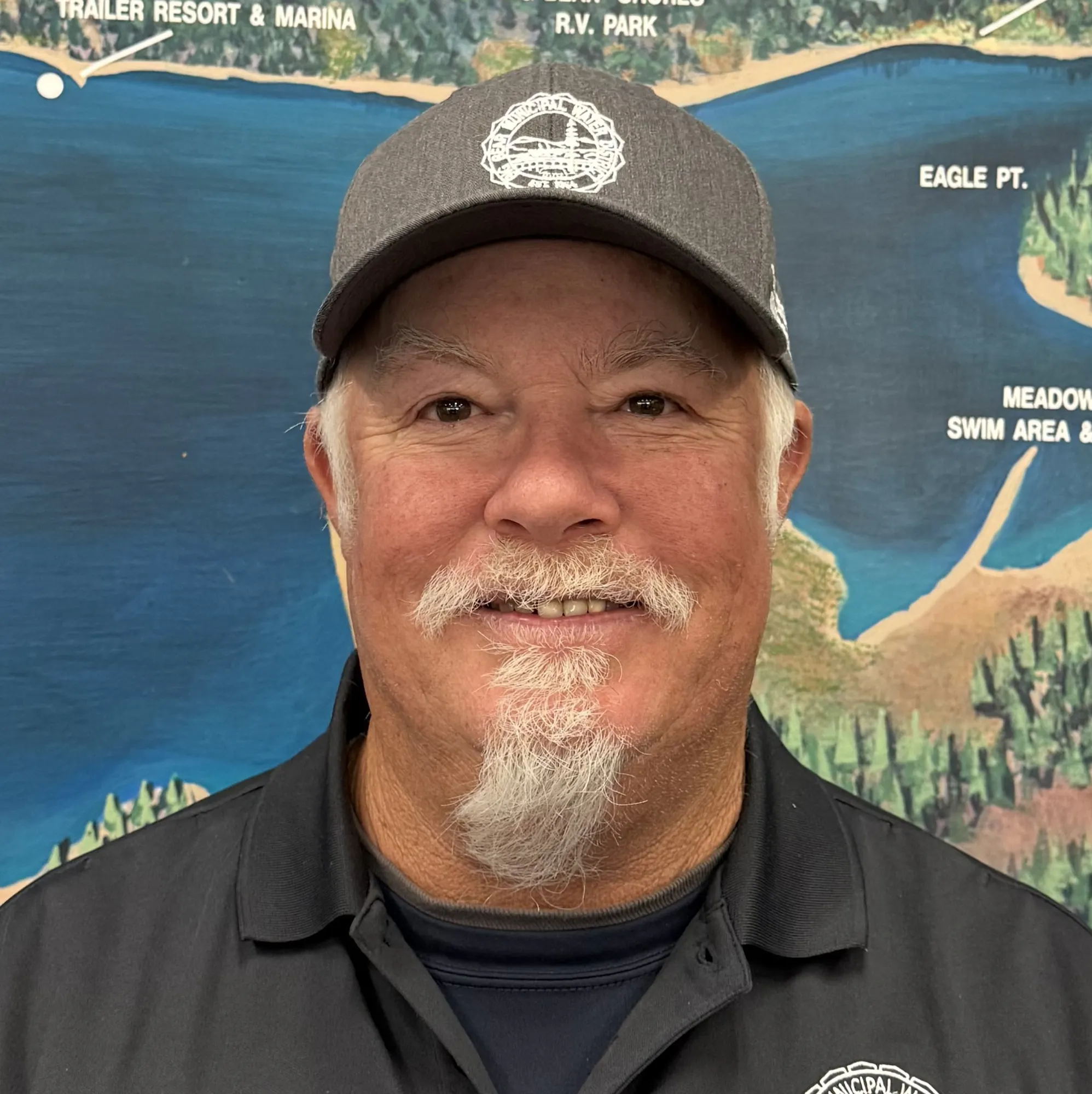 A man in a dark polo shirt smiling in front of a lake management map, wearing a gray cap, with the name "Peterson" on his shirt.
