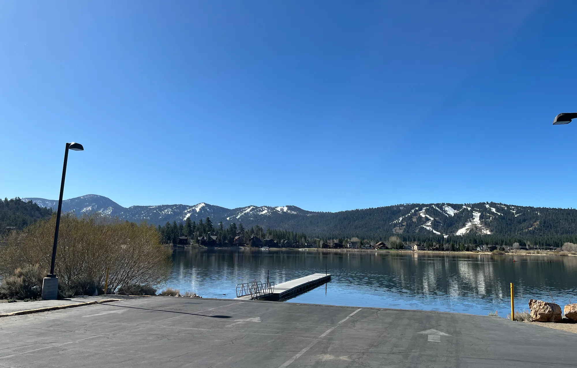 A serene lake with a dock, surrounded by mountains with patches of snow, under a clear blue sky.