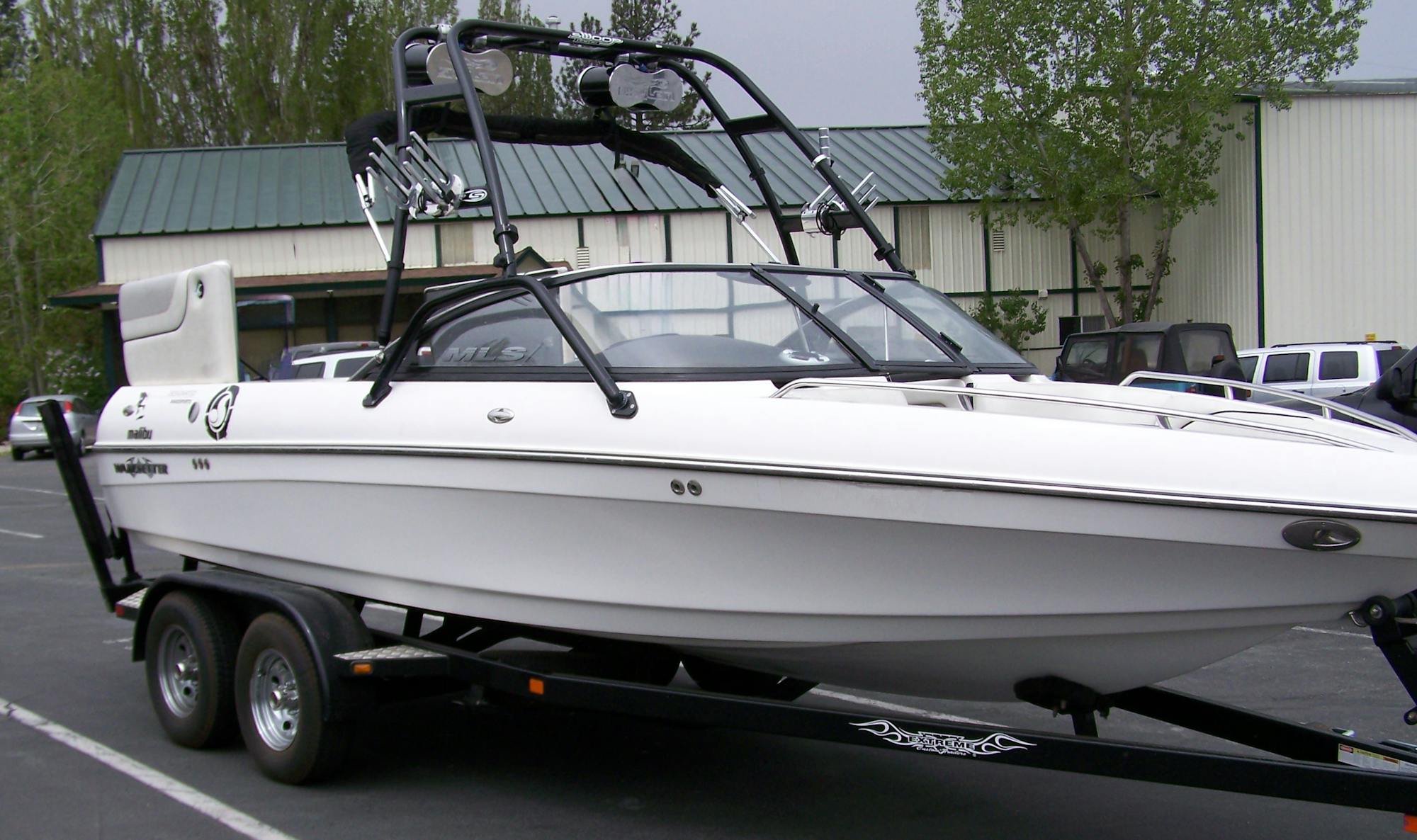 A white wakeboard style boat on a trailer parked in a parking lot.